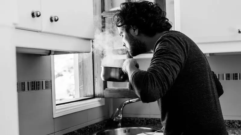 A younger man at his kitchen sink smells the steam wafting up from a pan