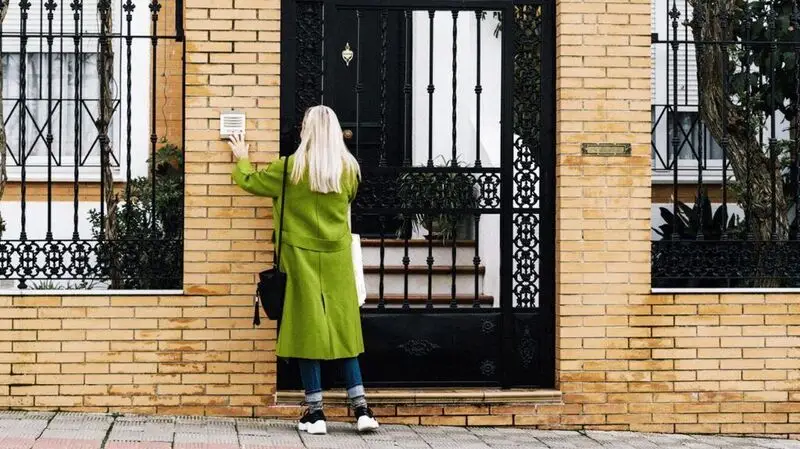 A blonde woman wearing a long green coat and sneakers rings the doorbell in front of a house