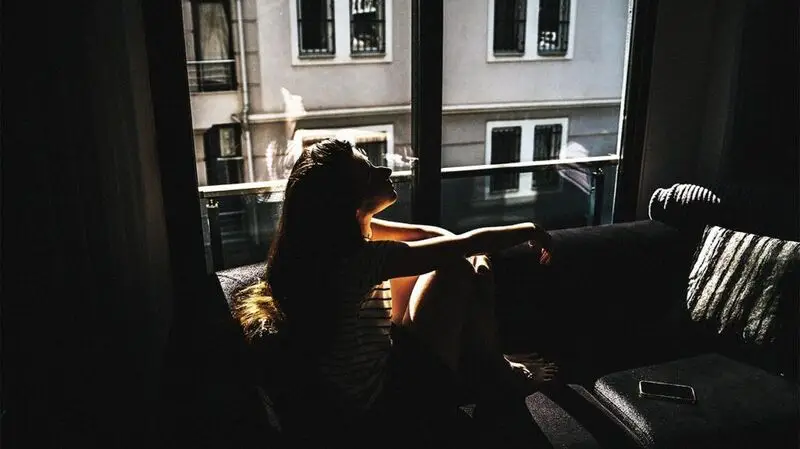 A younger woman sits in a darkened room near a window