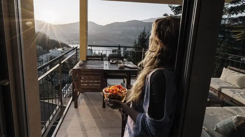 A woman holds a bowl of vegetables while looking at the scenery from a terrace