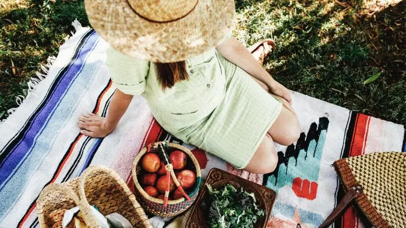 A pregnant woman sits on a blanket next to baskets of apples, nuts, and greens