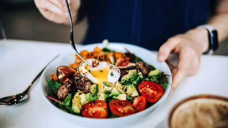 A person eating a plate of salad with vegetables, avocado and a egg on top
