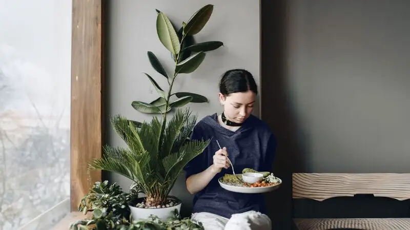 A woman eating a plate of food depicting a health meal plan while sitting next to a potted plant