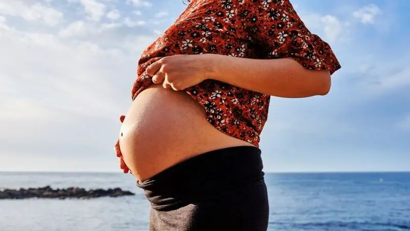 A woman shows her pregnant stomach while standing on a shore