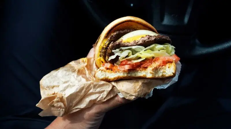 A person holds a double cheeseburger with onion, lettuce, and tomato