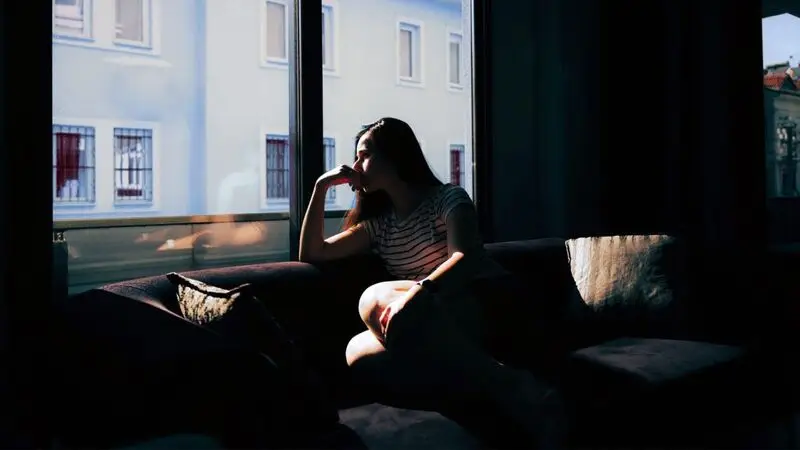 A woman looks out the window while sitting on a sofa in a dark room