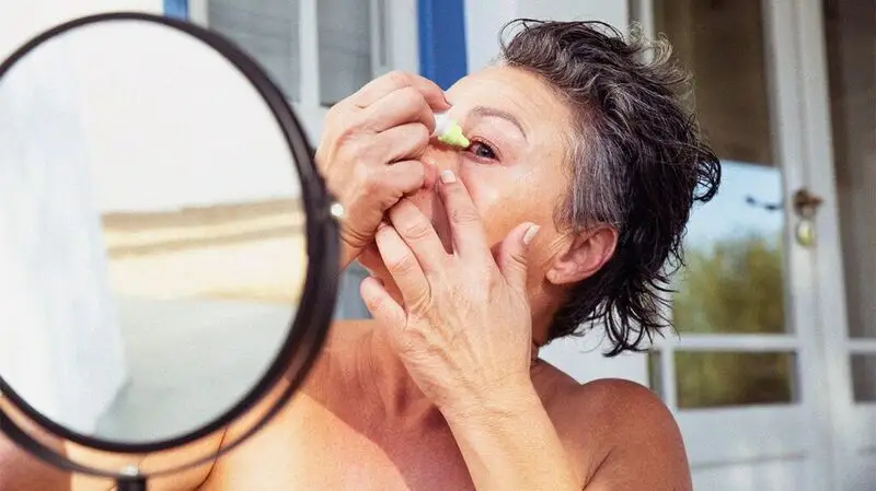 person with short black hair using eye drops in front of a round mirror