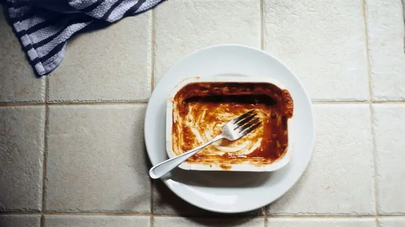 An empty microwave dinner container on a plate with a fork.