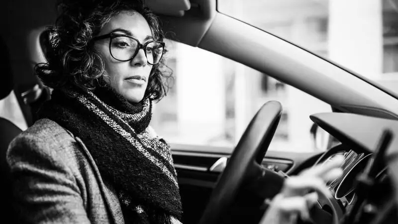 a woman wearing glasses and scarf sits in the driver's seat of a car