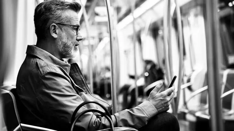 An older man wearing glasses and a jacket reads from his mobile phone while commuting on the subway