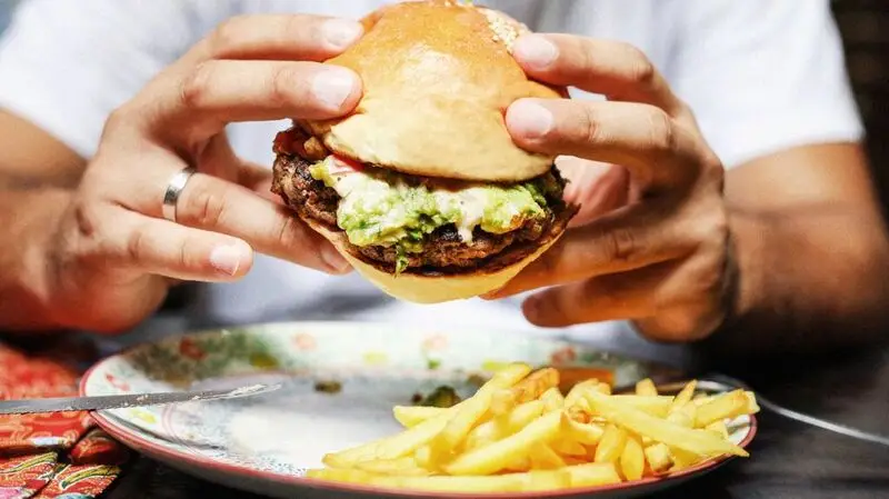 A person holding a cheeseburger above a plate of fries, both depicting high fat foods