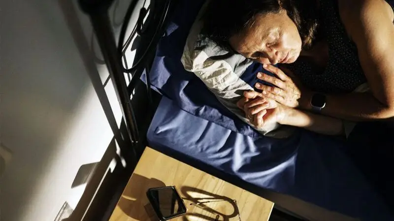 A person sleeping on a bed, turned towards their bedside table and the sun shining on them