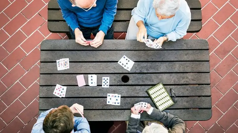 A bird's eye view of four older adults playing a game of cards on a wooden picnic bench outside