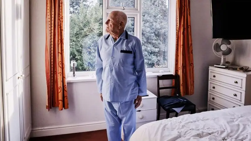 An older adult man stares out the window in a bedroom while wearing pyjamas