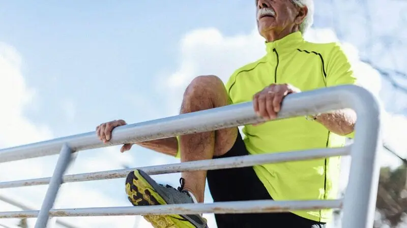 An older man in athleisure wear strecthes one leg high up on a railing