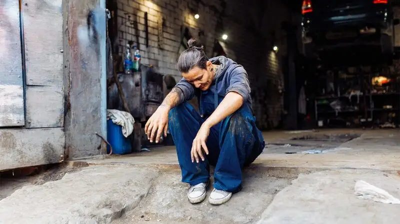 An older woman sits on the ground outside an auto shop while closing her eyes and resting her head on one arm