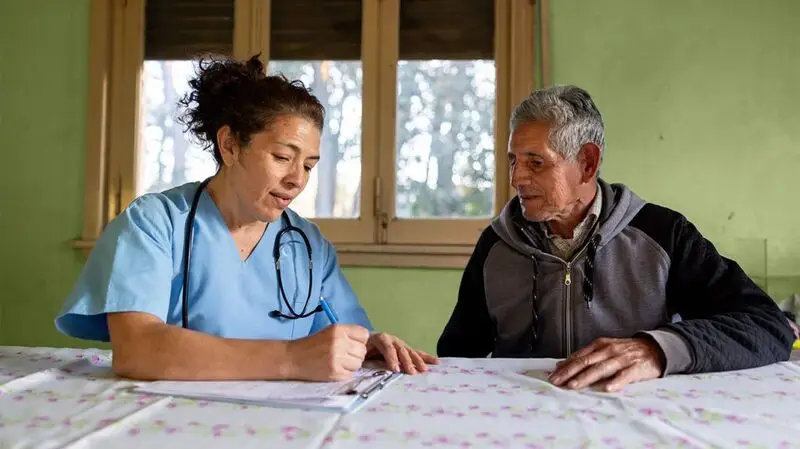 female doctor in blue scrubs speaking to older male patient in grey hoodie