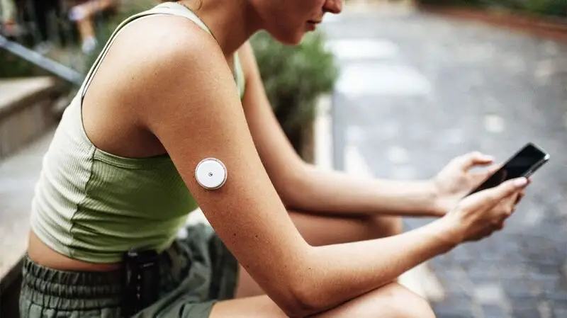 A woman in workout gear and wearable glucose monitor for diabetes sits to rest on a bench after a run