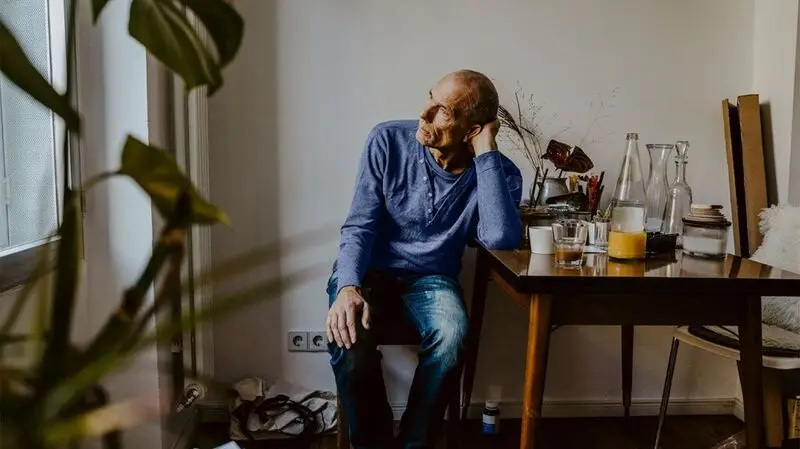 An older adult male sits at a table, resting his head in his head, staring pensively out of a window