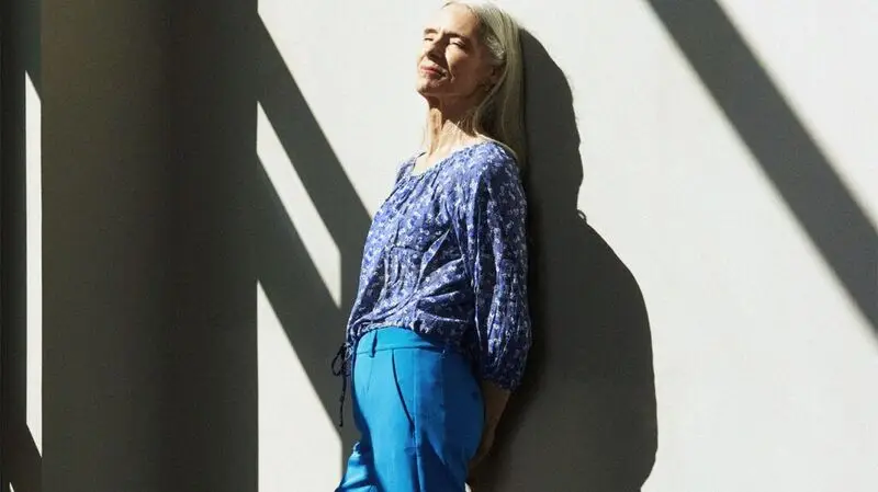 An older adult woman leans against a wall on a sunny day