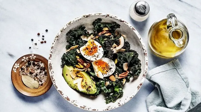 A bowl of kale salad with boiled eggs, nuts, and avocado on a white background