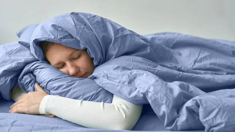 A person sleeping on a bed wrapped up in a duvet and hugging a pillow with their eyes closed