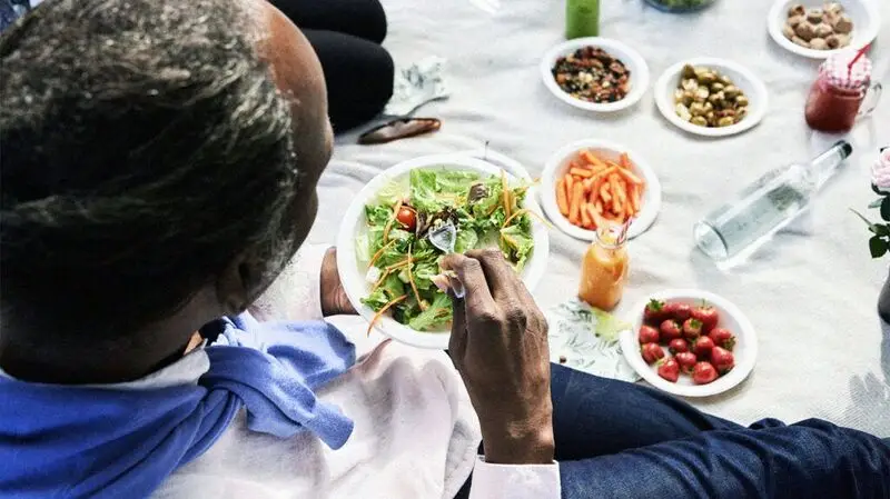 An adult eats a bowl of salad from a table full of plates of fruits and vegetables.