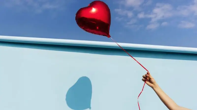 A person holding a heart shaped balloon against a backdrop featuring a wall and the blue sky