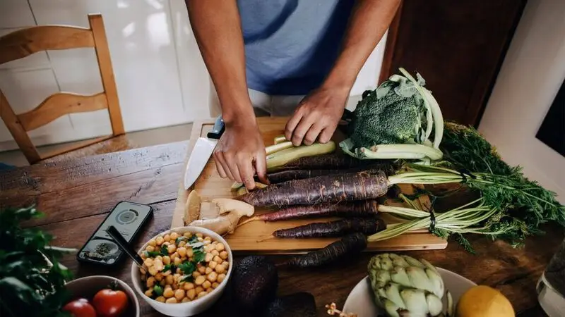 A perosn chopping various vegetables on a counter and chopping board, featuring purple carrots, broccoli, and artichokes, to showcase sources of luteolin