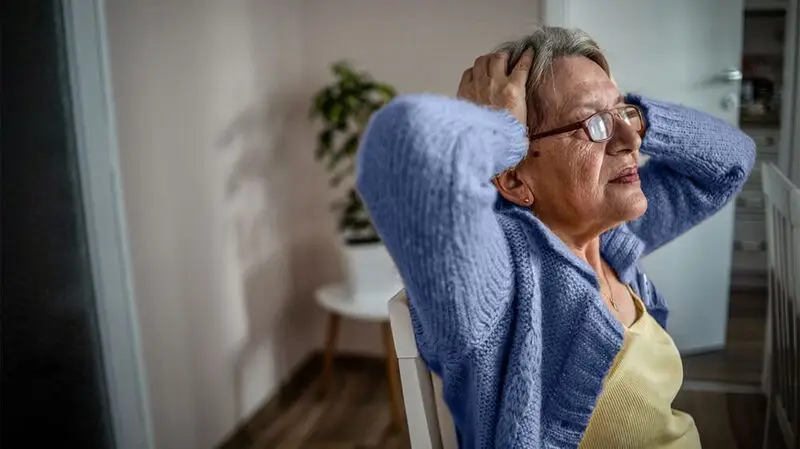 An older adult holds their head between their hands, elbows opened to the sides, while sitting in a chair indoors