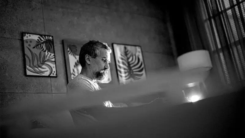 An older adult male sits in a living room with three paintings on the wall in the background