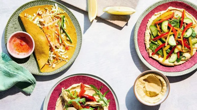 Three plates of plant-based foods, including tortillas full of vegetables, and a small bowl of hummus, and two lemon slices at the top