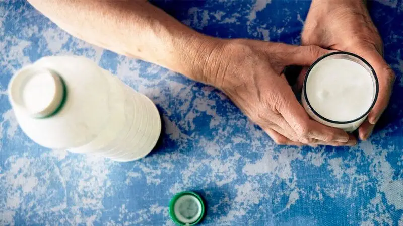 An older adult sits at the table with a glass of kefir in their hands next to an open bottle