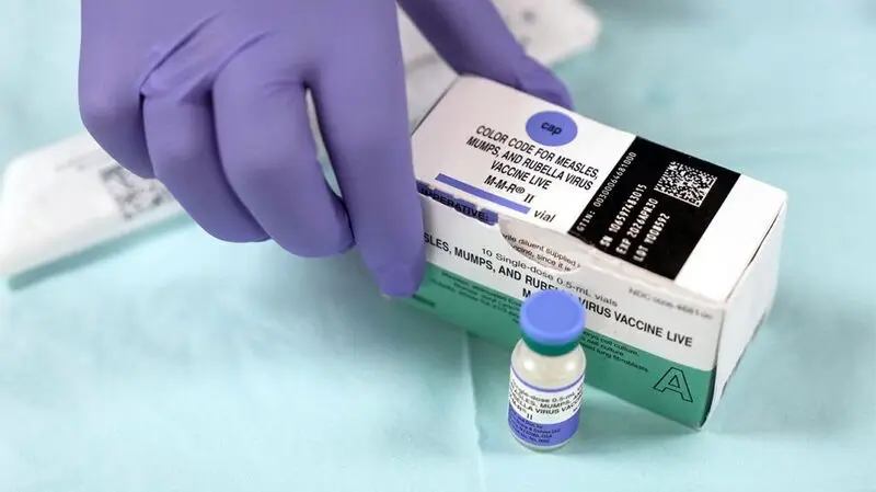 A gloved hand holds a box full of vials of the MMR vaccine against measles as a single vial is in front of the box