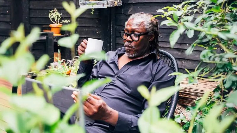 An older adult man sits on a chair outside with a cup in his hadn while looking at his tablet computer as the camera focuses on him through foliage