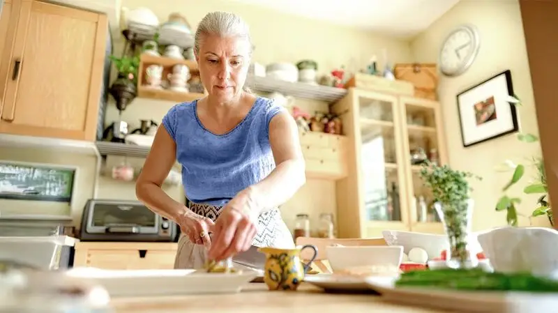 An older adult woman preparing a Mediterranean salad in a kitchen with a knife, chopping board and various utensils