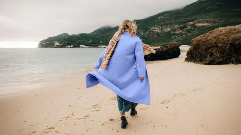 A woman walks on the beach as her long jacket floats in the wind