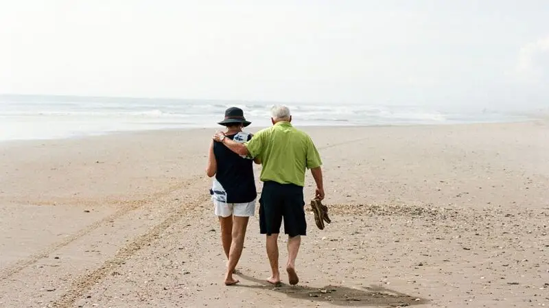 Two older adults walking on the beach barefoot as one puts an arm over the other and holds their shoes in the other hand