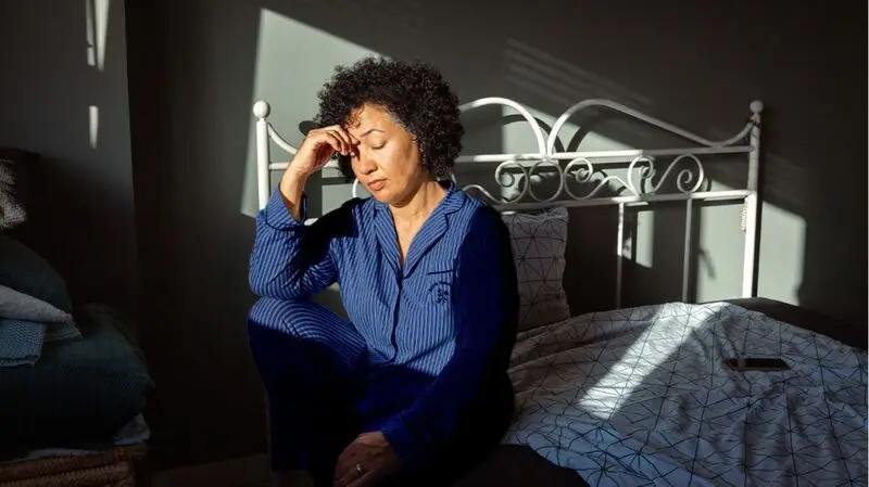 A woman hold her eyebrow, resting her face in her hands, looking tired on her bed