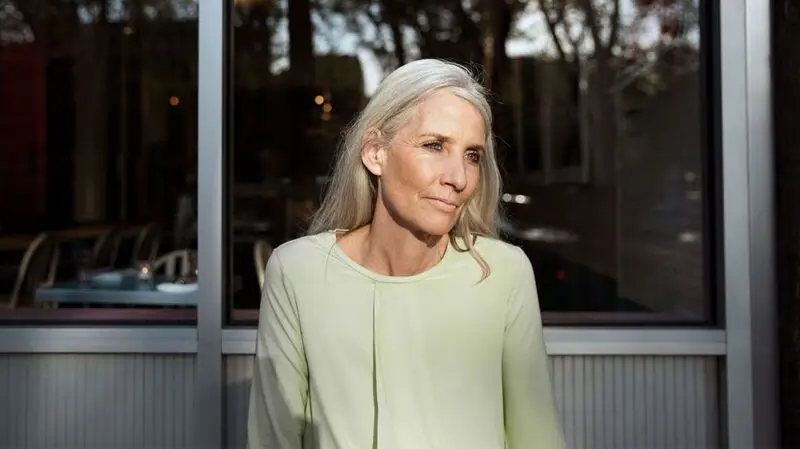 An older adult woman looks slightly to the side, outside as the sun shines on her in front of a glass panel building