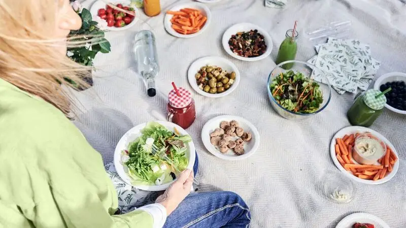 A person sits on a picnic blanket with various plates of vegetables, fruits, and nuts on plates that show a healthful diet