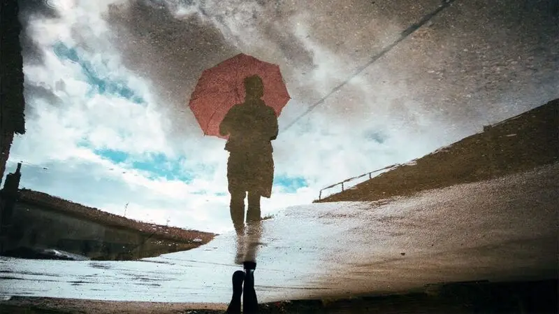 A person walks in the rain with an umbrella as their reflection shows up in a puddle on the ground