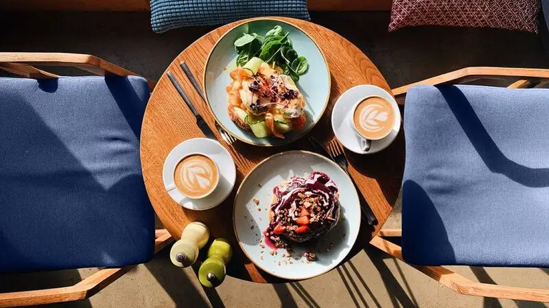 An overhead view of a breakfast table feaurting a plate of green salad, salmona nd avocado toast, pancakes and fruit, and two cups of coffee