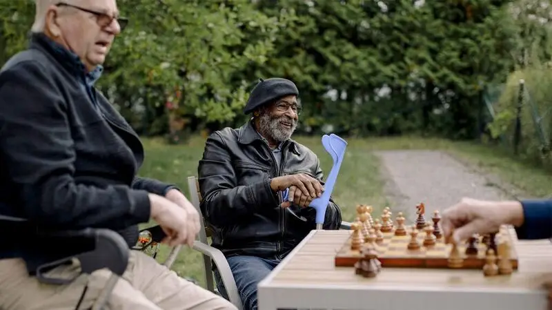 Three older adult men play chess outside, as one of them watches the other two playing, his hands resting on a walking aid