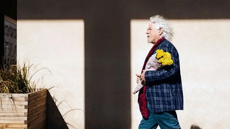 An older man wearing a blzer and scar, holding a bouquet of flowers walks outside during a sunny day, AS his hair IS swept away by the wind