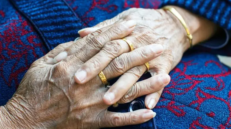 A close-up of an older person's hands, fingers interwoven, wearing gold rings and a gold bracelet