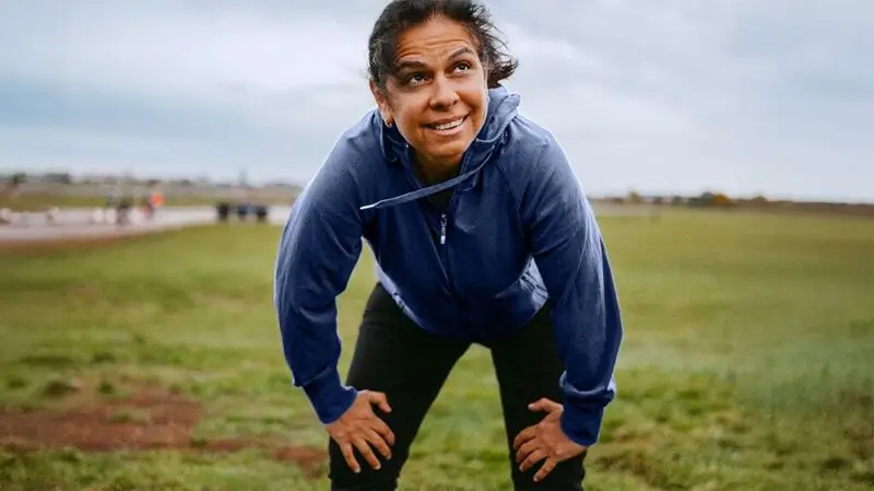 A middle aged woman holds her knees, her body slightly bent over, catching her breath after a run outside