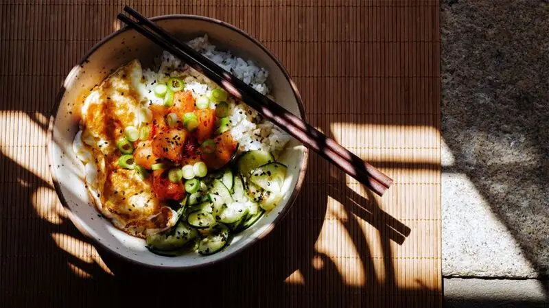 chopsticks resting on bowl of fish, rice, egg, and cucumber slices