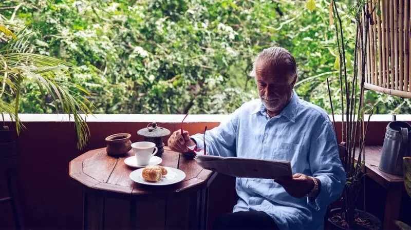 An older adult reads a newspaper with his glasses in hand as he has a breakfast of coffee and a croissant next to a table near a window
