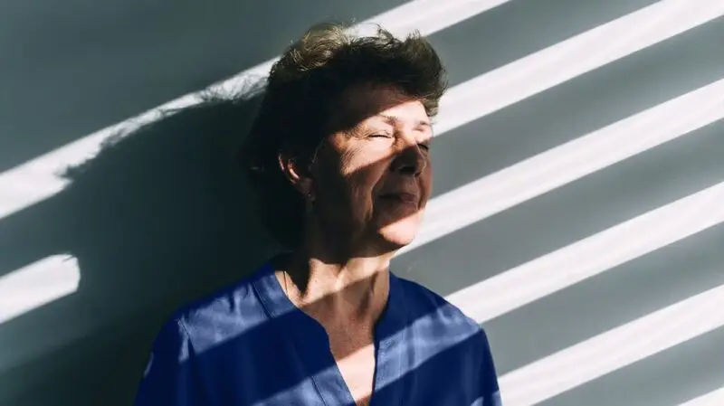 A woman leans against a wall with her eyes closed as the shadows from a window blind reflect sunlight on her face